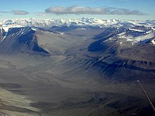 In the foreground is a landscape of dull brown mounds and undulations, behind which are snow-covered mountain peaks.